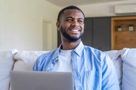 Young man smiling while working on a laptop at home on a sofa, relaxed remote worker enjoying comfortable modern living room.