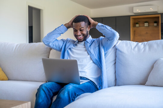 Young man sitting on a couch at home with a laptop, smiling and surprised while working remotely, celebrating good news and enjoying casual relaxed remote work in a bright living room