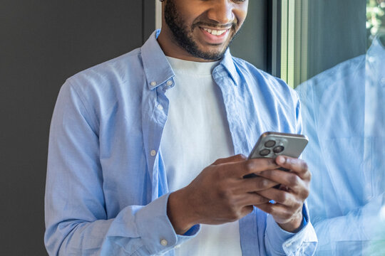 Smiling young man using smartphone and texting by a window, casual lifestyle portrait showing mobile technology, connection, and relaxed communication