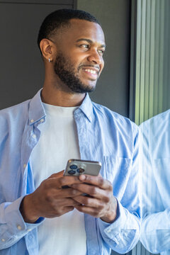 Young man smiling and holding smartphone while looking out a window, wearing a casual shirt; relaxed and thoughtful mood suggesting connection, communication, remote work and modern lifestyle