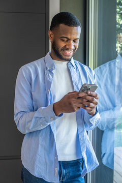 Young man smiling and texting on a smartphone while standing by a window in a casual shirt, using mobile phone for messaging, social media and communication at home or office