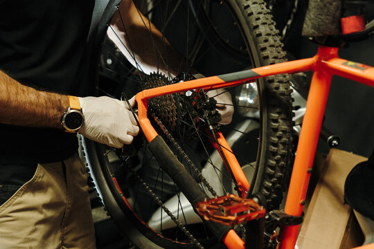 Male bicycle mechanic services the rear drivetrain of an orange mountain bike. Gloved hands work around the cassette, chain and rear wheel during maintenance
