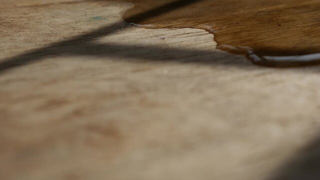 Macro close-up of thin water stream trickling across rustic aged wood grain surface with warm golden light and shadow in still life photography