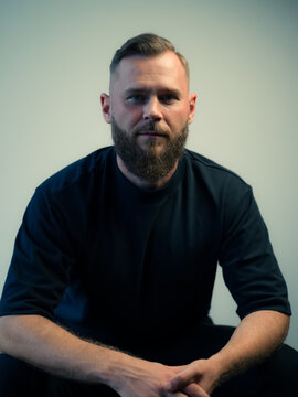 Man With Beard Sits Calmly Against a Plain Background
