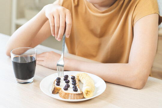 Diet concept asian young woman, girl having a healthy breakfast with banana and blueberry toast with cream cheese on a white plate, served with hot black coffee on a wooden table in the morning light