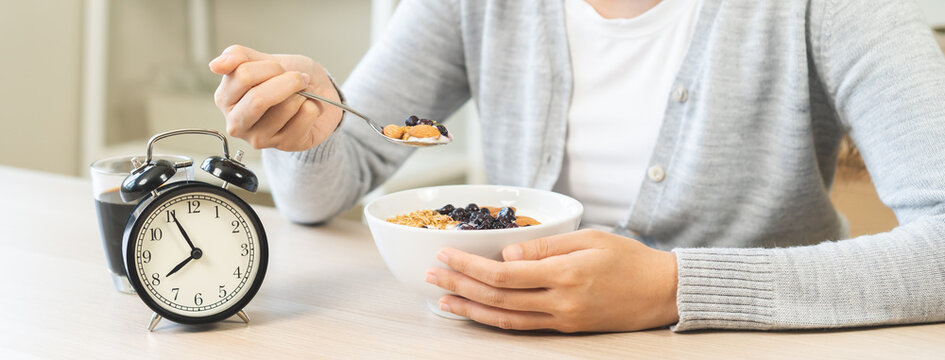 Young woman eating a healthy Greek yogurt bowl with granola and blueberries at a wooden table in the morning, with alarm clock showing the breakfast time.