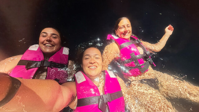 Three women smiling and wearing life jackets swimming at night