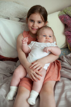 A young girl holds her newborn sibling on a soft bed at home