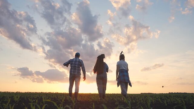 A man, a woman, and a girl walk through the field at sunset. The man, the woman, and the girl carry tools and gaze at the beautiful sky.