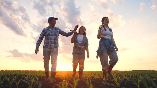 A man, a girl, and a woman walk together through a field as the sun sets. They carry gardening tools, sharing smiles against a vibrant sky.