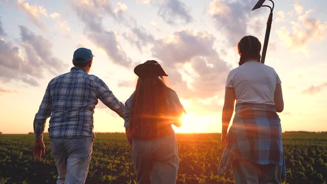 A man, woman, and girl walk together in a field. The man gestures towards the sunset while holding gardening tools, enjoying the evening sky.