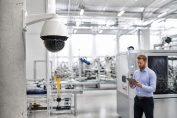 Fototapeta premium A security camera is mounted on a pillar in a bright, modern factory setting where a man is looking at a tablet.
