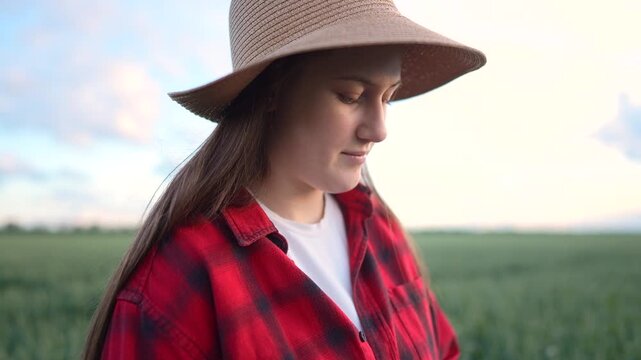 The girl in a straw hat stands in a field, the girl observing the horizon with a contemplative expression. The girl wears a red plaid shirt over a white t-shirt.