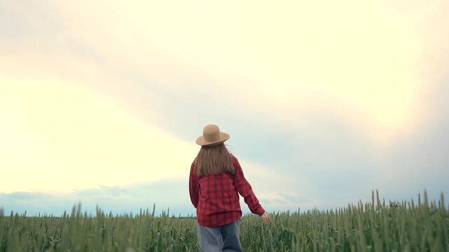 A woman in a red flannel shirt walks through a field of green grass, with a straw hat shading her face under the expansive sky. The woman enjoys the serene landscape while strolling thoughtfully.