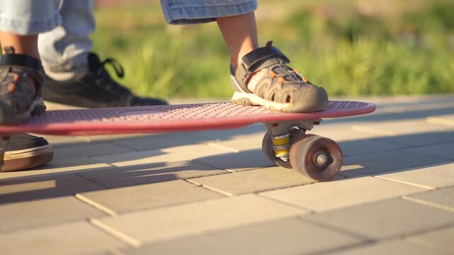A child balances on a skateboard while an adult supports the child. The child wears sports shoes and denim pants, practicing skateboarding outdoors.