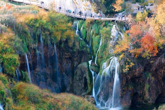 A gorgeous high-angle vista captures the majestic Milanovac Waterfall pouring into the turquoise pools of Plitvice Lakes, bordered by a vibrant autumn forest and a crowded cliffside boardwalk