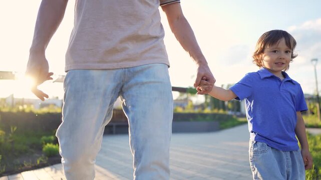 A man and a boy walk hand in hand along a pathway. The man and boy enjoy their time outdoors together, with smiles on their faces as sunlight shines.