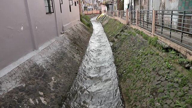 Flowing water rushes through narrow concrete channel. Wet stone walls frame urban drainage path. Weathered buildings line both sides of the stream. Green moss grows along cracked concrete edges