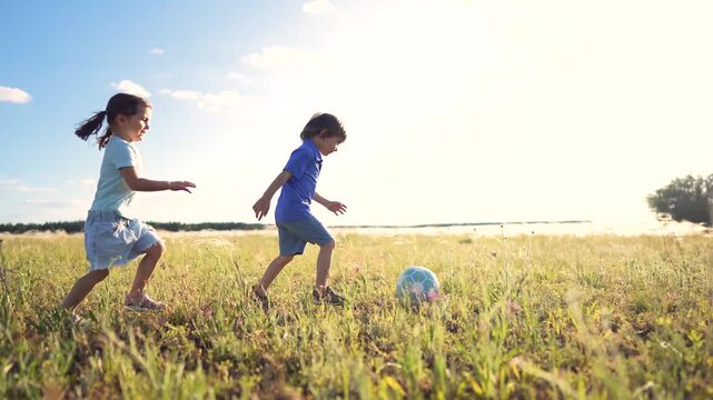 A girl runs joyfully while a boy plays soccer in a field. The boy and girl enjoy the sunny day as they kick the soccer ball in the green grass.