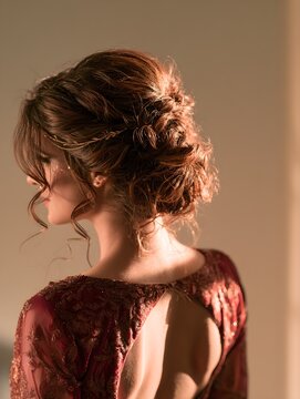 Elegant woman with intricate messy updo hairstyle wearing luxurious maroon embroidered evening dress standing under warm glowing soft studio lighting