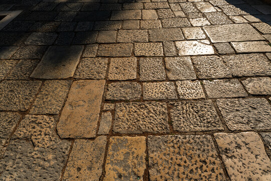 Weathered cobblestone pavement with reflective wet stones and long shadow across the surface
