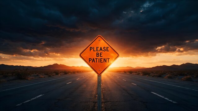 Bright orange diamond shaped road sign displaying please be patient text standing in middle of long dark empty asphalt highway during golden sunset evening light