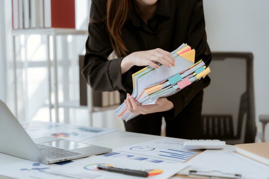 Organized Workflow: A professional woman meticulously sorts through a stack of documents, her desk adorned with charts and a laptop, embodying efficiency and order in a modern office setting.