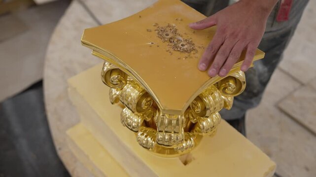 Hand using power drill on gilded classical column capital. Warm golden tones contrast with muted workshop background