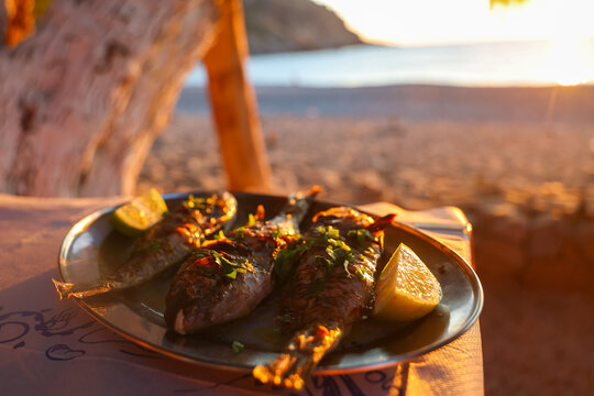 A plate of grilled fish garnished with herbs and lemon sits on a seaside table at sunset, capturing Mediterranean cuisine and coastal dining atmosphere.