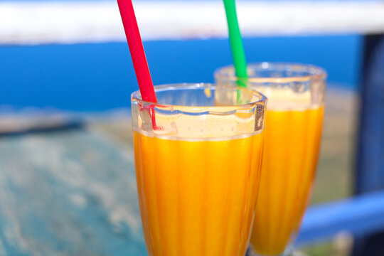 Two tall glasses of bright orange mango smoothies with colorful straws rest on a wooden table overlooking the vivid blue sea under clear summer skies.