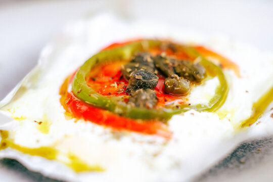 A traditional Greek baked cheese dish topped with peppers, tomatoes, capers, and olive oil glistens in sunlight, representing Mediterranean flavor and culinary heritage.