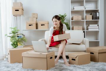 Fototapeta premium Woman Preparing Packages for Shipping: A young woman sits in a well-lit room surrounded by cardboard boxes, laptop open on her lap, as she meticulously prepares items for shipment.