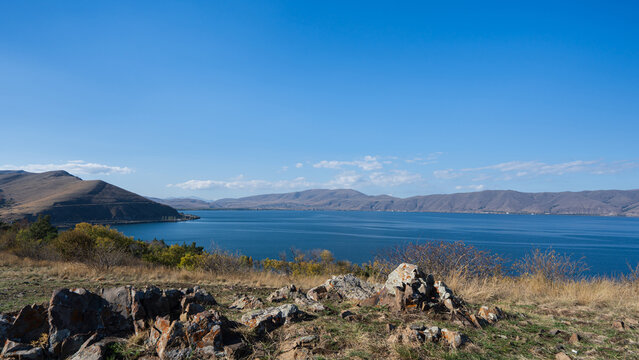 View of Lake Sevan framed by lichen-covered rocks and autumn grassland, with mountains under a clear blue sky, Armenia.