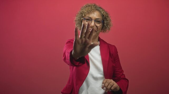 Woman with curly hair places palm to camera in studio, wearing red blazer and glasses; confidence boundary.