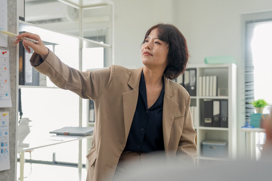 Confident businesswoman pointing at whiteboard chart, explaining strategy and presenting data during office meeting, close-up dynamic angle capturing leadership and business presentation concept