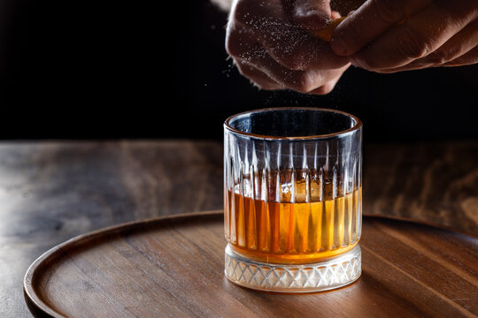 Hands pressing an orange peel releasing the oils on top of an old fashioned cocktail on a wooden bar top.