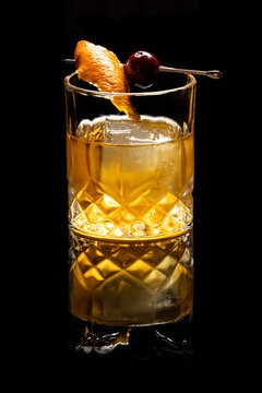 Close up of a classic old fashioned cocktail on a dark reflective background garnished with a cherry and an orange twist.