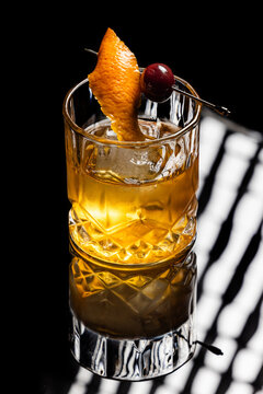 Close up of a classic old fashioned cocktail on a dark reflective background garnished with a cherry and an orange twist.