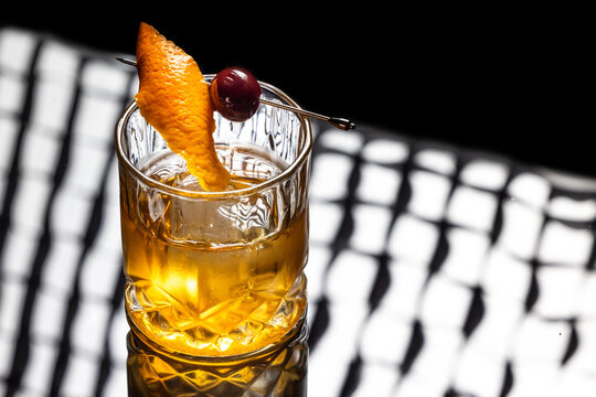 Close up of a classic old fashioned cocktail on a dark reflective background garnished with a cherry and an orange twist.