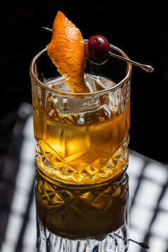 Close up of a classic old fashioned cocktail on a dark reflective background garnished with a cherry and an orange twist.
