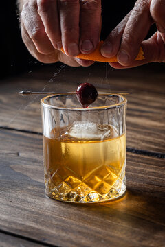 Hands pressing an orange peel releasing the oils on top of an old fashioned cocktail on a wooden bar top.
