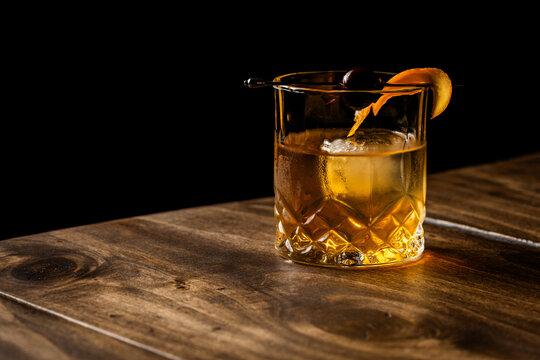 Close up of a classic old fashioned cocktail on a wooden background garnished with an orange twist and a cherry.