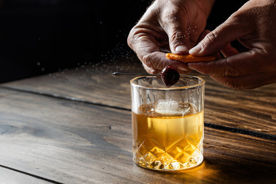 Hands pressing an orange peel releasing the oils on top of an old fashioned cocktail on a wooden bar top.