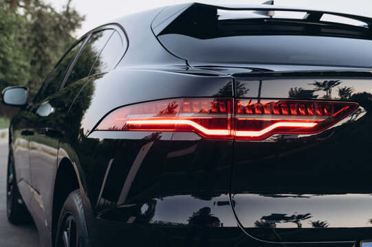 Close up of a black car rear with an illuminated red taillight, sleek bodywork, reflective surface, and rear spoiler in soft evening light.