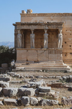 The Porch of the Caryatids at the Erechtheion temple rises above scattered ancient stones on the Acropolis of Athens, framed by clear blue sky and distant mountains.