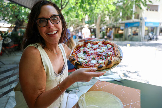 A smiling woman holds a freshly baked pizza topped with tomato, mozzarella, and basil at an outdoor caf&eacute;, enjoying Mediterranean cuisine and summer leisure in Athens.