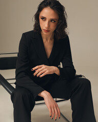 Profile portrait of an elegant young woman with short wavy hair and makeup, wearing a black suit sitting on a chair in a studio on a neutral gray background. © Corina