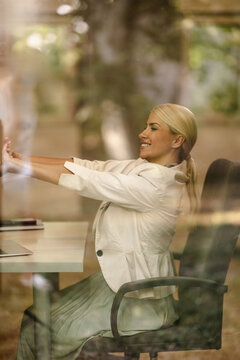 Woman Works in a Small Office During the Day, Stretching and Smiling Through the Glass