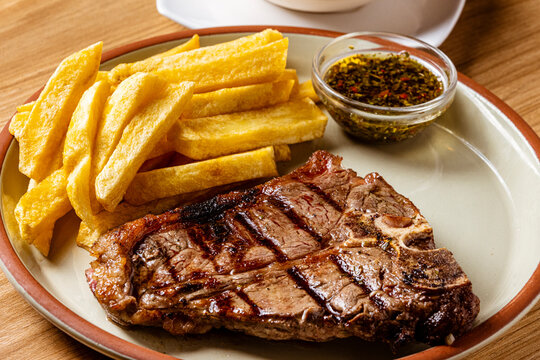 Grilled T-Bone Steak Served with Crispy French Fries and Chimichurri Herb Sauce on Ceramic Plate