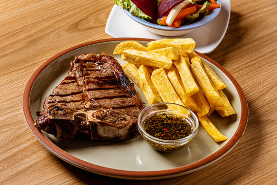 Grilled T-Bone Steak Served with Crispy French Fries and Chimichurri Herb Sauce on Ceramic Plate
